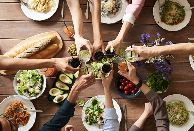 A group of people seated around a dining table clinking glasses in a toast. The table is set with various dishes, including salads, pasta, bread, and avocado.