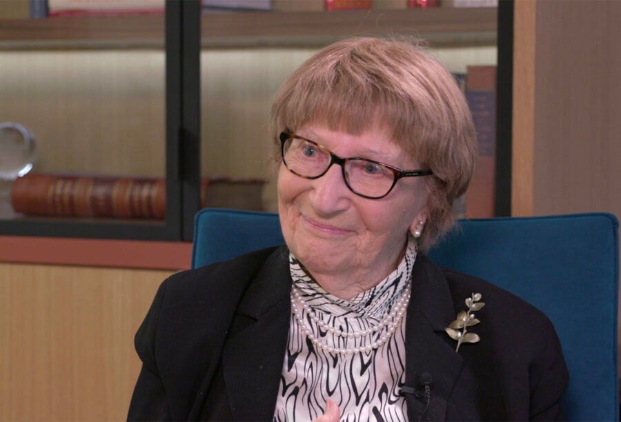 An elderly person with short hair and glasses, wearing a patterned blouse and a black jacket with a leaf-shaped brooch, is sitting in a room with bookshelves.