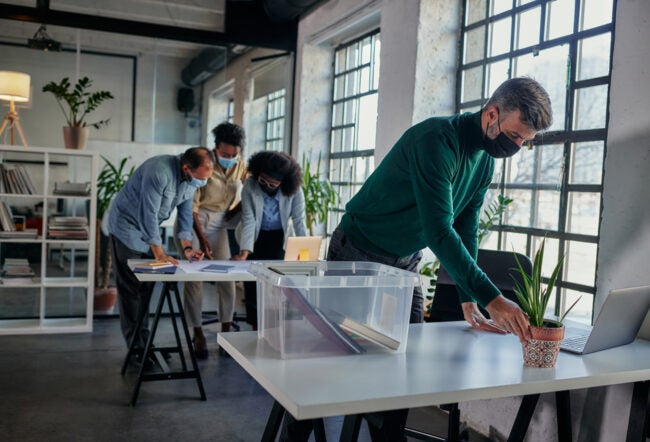 People wearing masks working in a bright office space with large windows. One person is arranging items on a table while others are huddled around a desk.