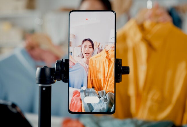 A smartphone on a tripod with a person live streaming, holding up orange and blue shirts, representing online shopping or fashion content creation.