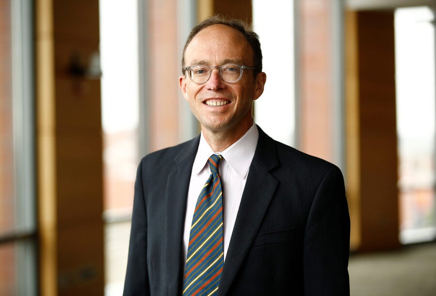 A headshot of a person wearing glasses, a dark suit, a white shirt, and a striped tie, standing in a well-lit indoor setting.