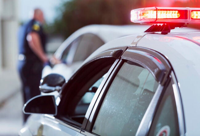 A police officer is talking to a person in a white car during a traffic stop. A police car with flashing lights is visible in the foreground.