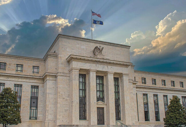A stately government building with a classical facade and a flagpole with two flags flying. Sun rays and clouds are visible in the sky above.